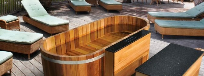 Drewniana wanna z hydromasażem na tarasie, otoczona wiklinowymi leżakami z jasnymi poduszkami.