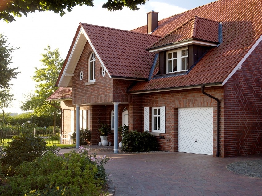 Dwukondygnacyjny dom z czerwonej cegły z garażem i czerwonym dachem, otoczony zielenią i kwiatami, widok z brukowanego podjazdu.
