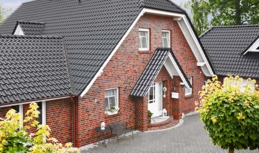 Dom z czerwonej cegły z czarnym dachem z dachówki karpiówki, białe obramowania okien i drzwi, widok z zewnątrz na podjazd z kostki brukowej.