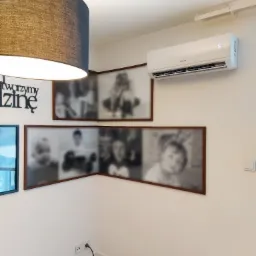 Biały klimatyzator ścienny zamontowany w rogu pokoju, pod nim trzy oprawione zdjęcia rodzinne, obok widoczny włącznik światła i lampa wisząca z abażurem.