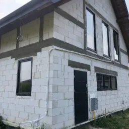 Surowy stan budynku z betonu komórkowego, z zamontowanymi oknami w czarnych ramach i czarnymi drzwiami wejściowymi, widoczne białe rury instalacji zewnętrznej, szare elementy wykończeniowe pod dachem.