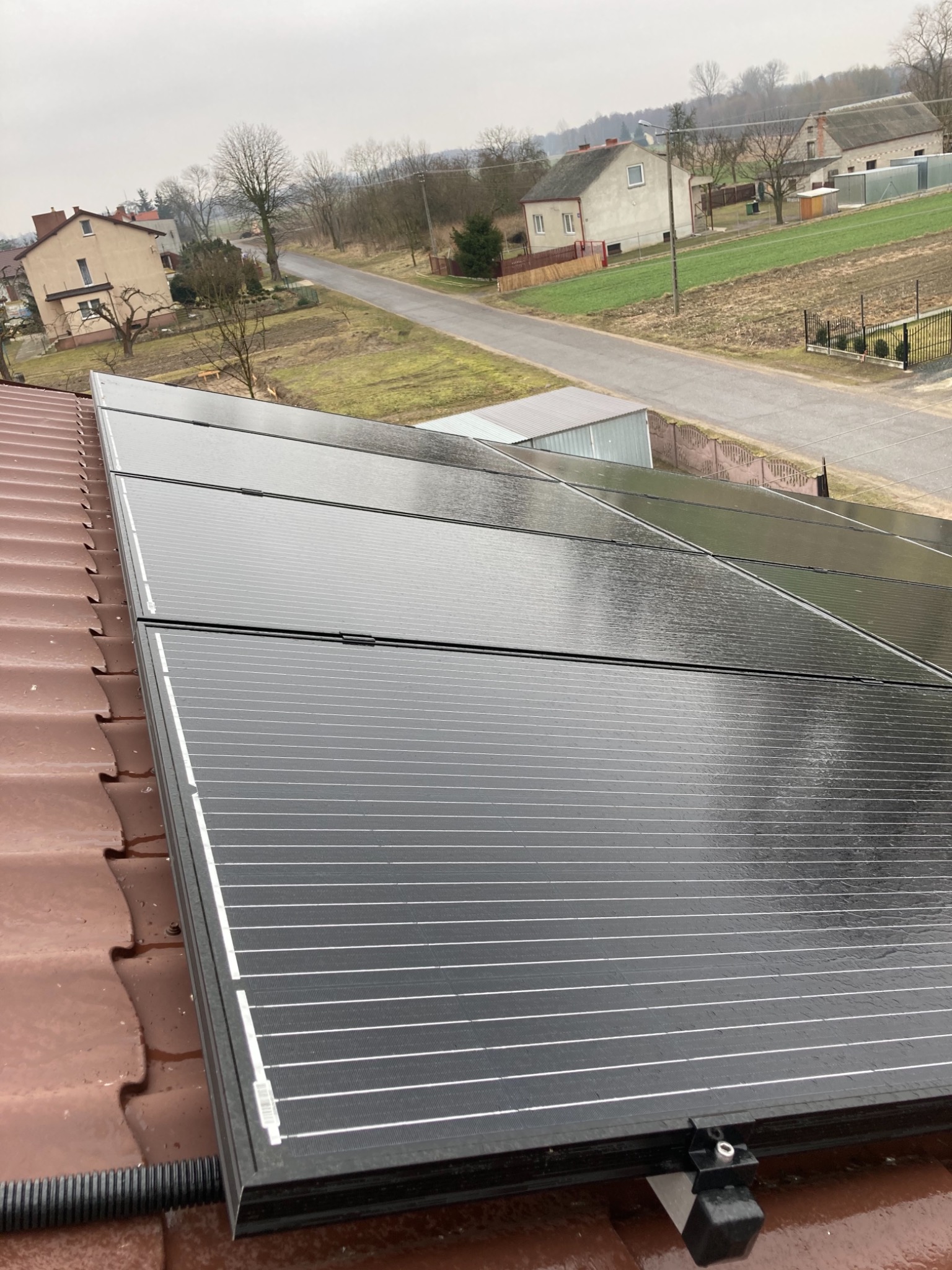 Panele słoneczne na brązowym dachu z widokiem na wiejski krajobraz w pochmurny dzień, z widoczną rurą karbowaną odprowadzającą przewody.