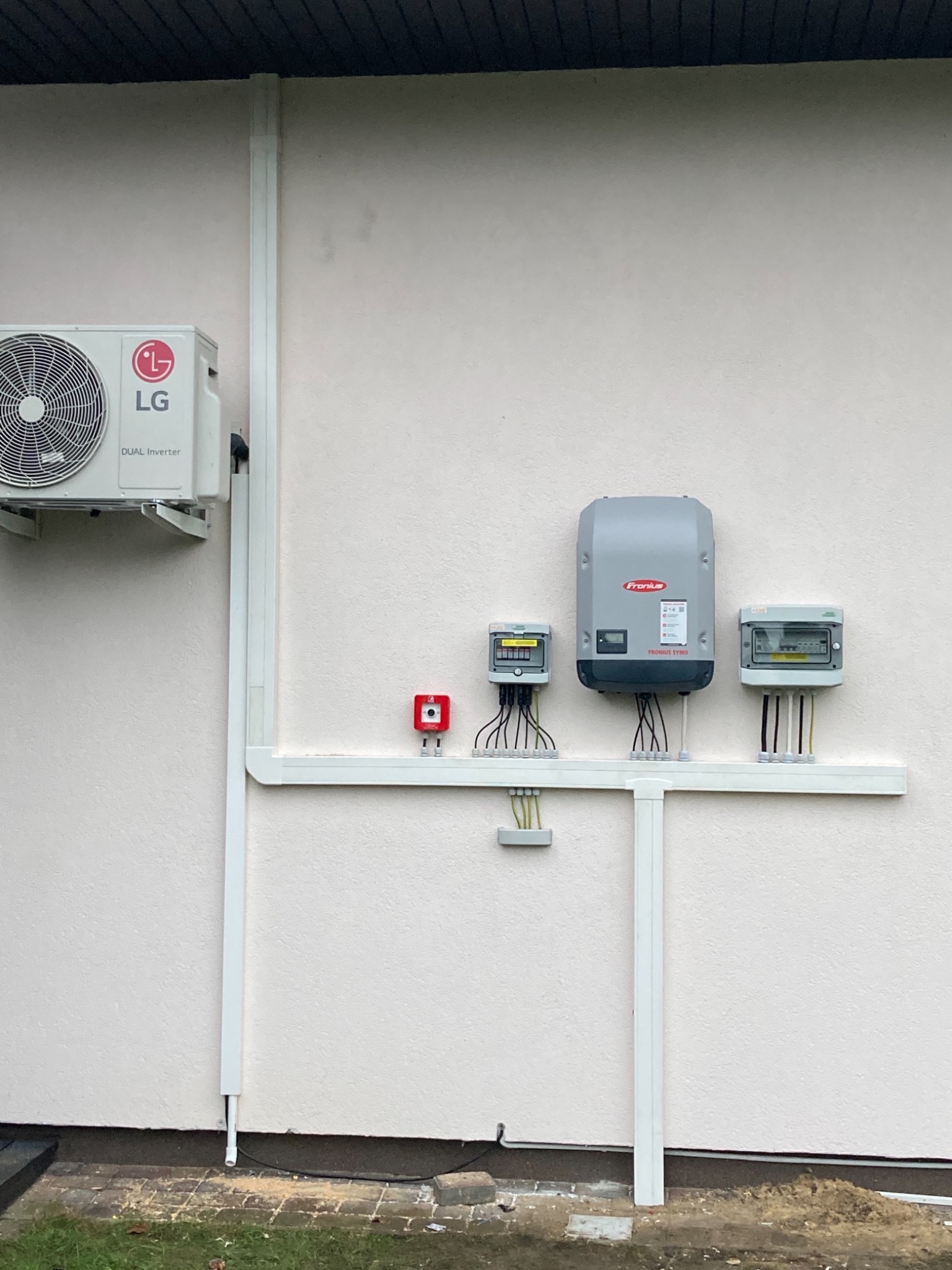 Inwerter Fronius Symo i skrzynki elektryczne zamontowane na elewacji budynku, z widocznymi przewodami w białych listwach, obok klimatyzator LG Dual Inverter.