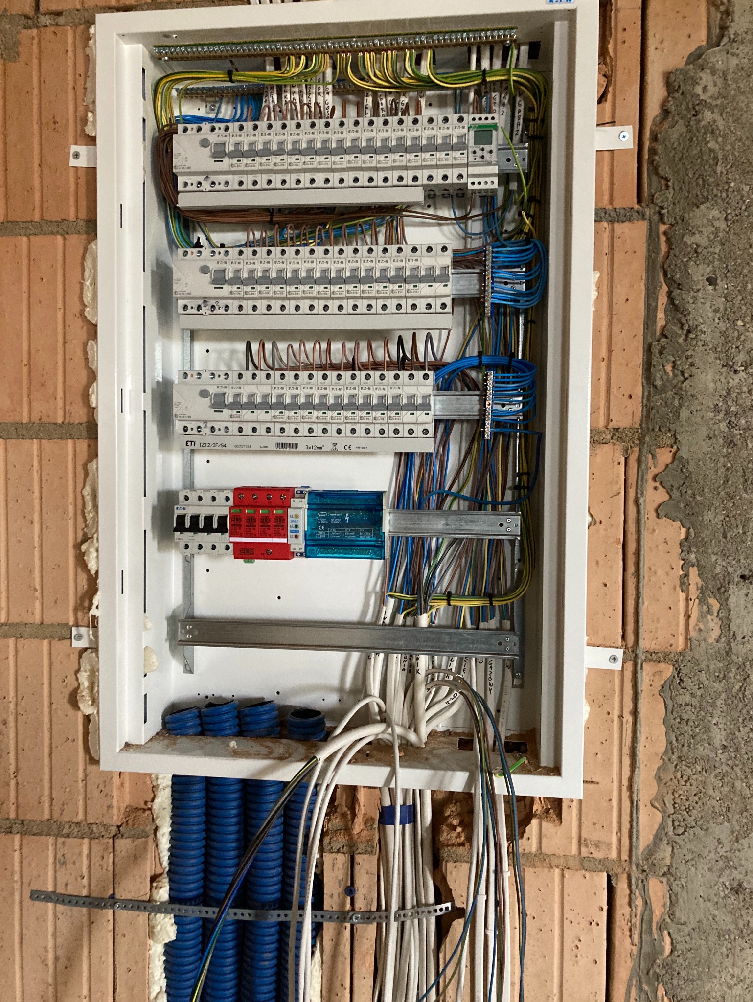 Rozdzielnica elektryczna w białej obudowie, zamontowana na ścianie z cegły, widoczne liczne przewody, zabezpieczenia ETI oraz niebieskie rury karbowane.
