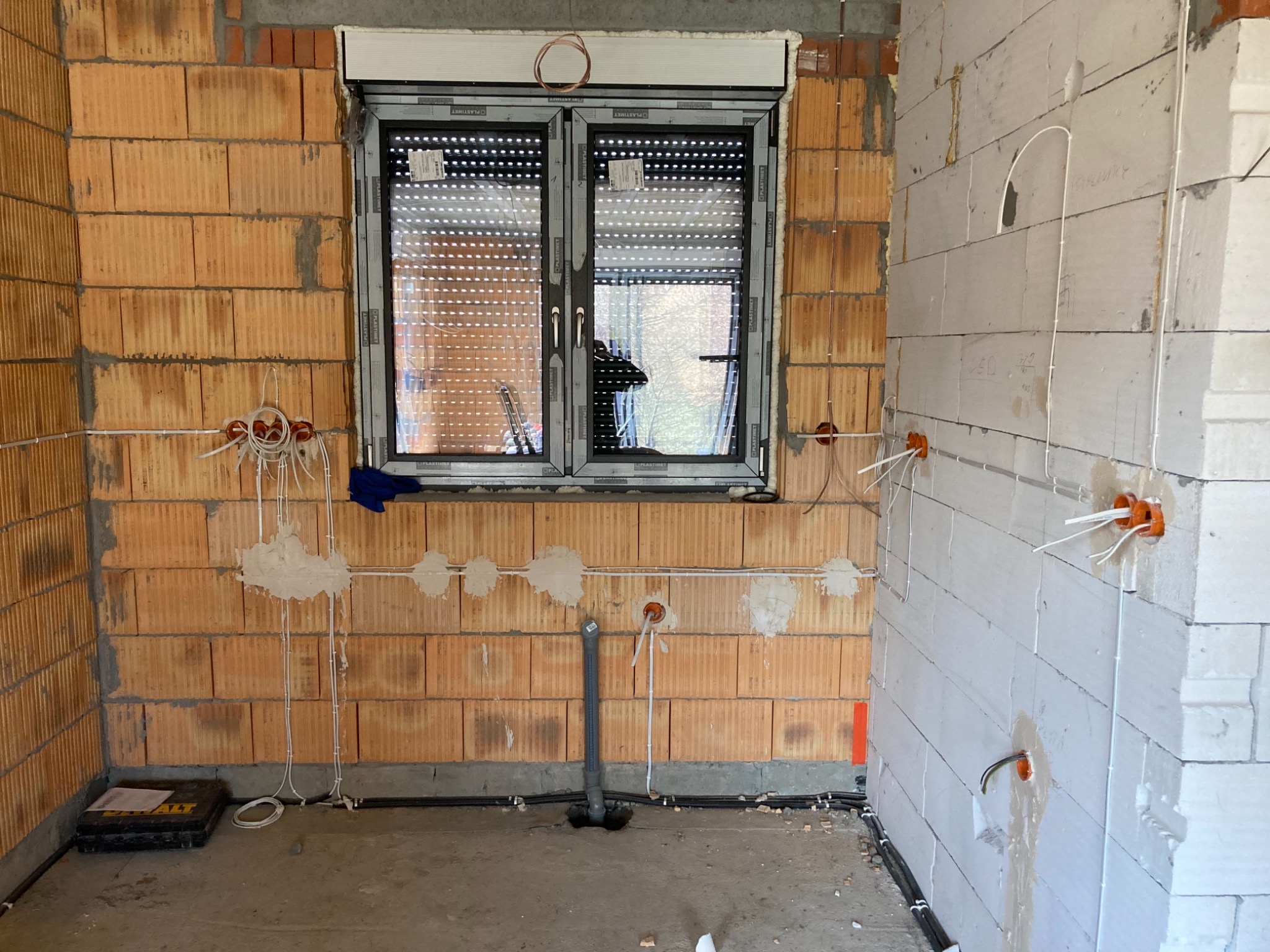Wnętrze w trakcie prac wykończeniowych: widoczne kable elektryczne wystające ze ścian z cegły i bloczków, puszki instalacyjne, okno z roletami zewnętrznymi i rura kanalizacyjna wystająca z podłogi.