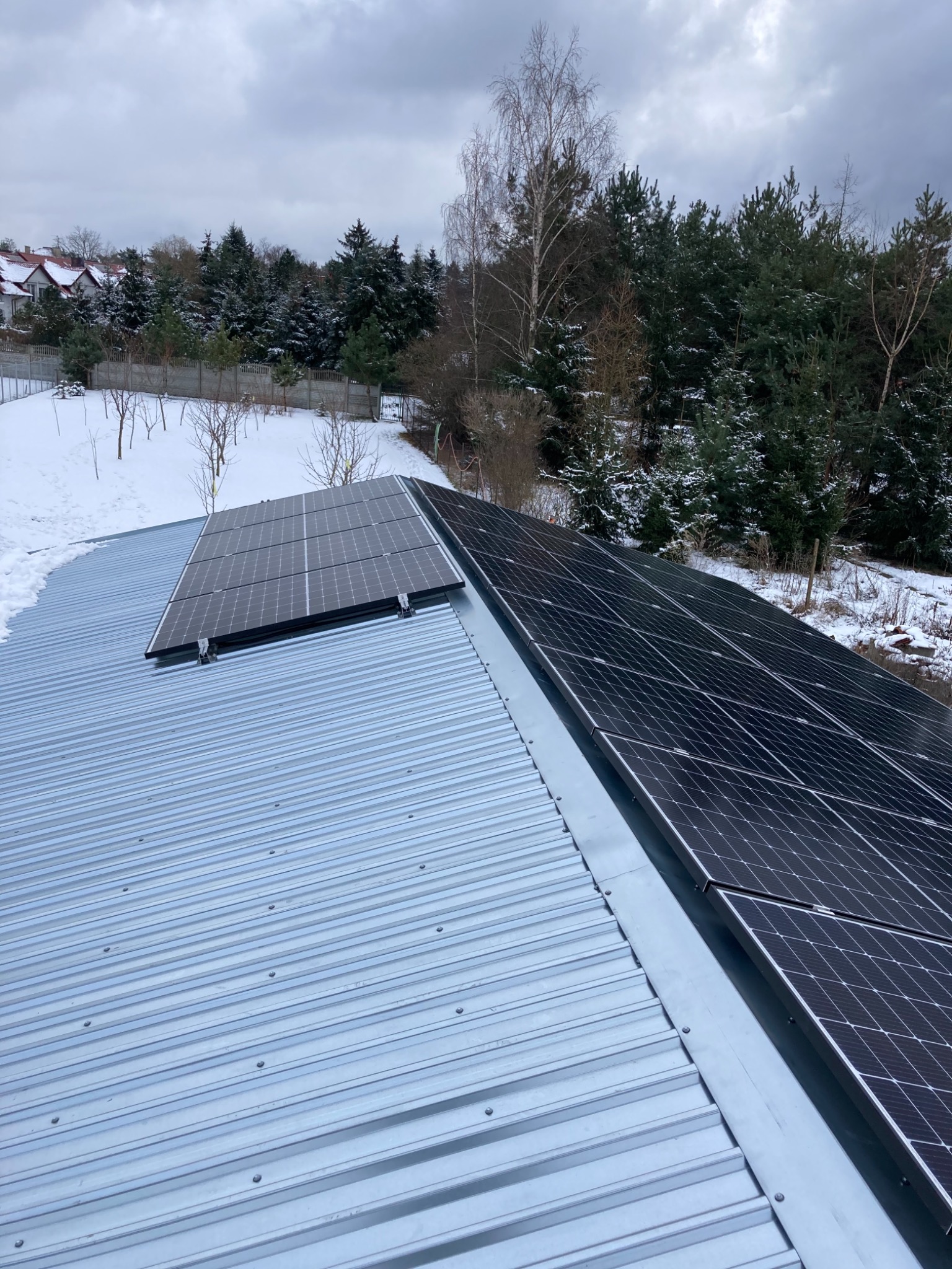 Panele fotowoltaiczne zamontowane na blaszanym dachu w zimowej scenerii, widok z góry na częściowo ośnieżone otoczenie.