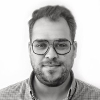 Portret mężczyzny w okularach z metalowymi oprawkami, ubranego w koszulę w drobną kratę, na jednolitym, jasnym tle. Zdjęcie czarno-białe.