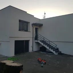 Nowoczesny dom z jasną elewacją, zewnętrznymi schodami prowadzącymi do drzwi wejściowych na piętrze, narzędzia budowlane i pniak drzewa na brukowanym podwórku.