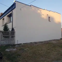 Boczna ściana domu po tynkowaniu i malowaniu na jasny kolor, widoczna mała kwadratowa okienko, ciemna podbitka dachowa i palety pod ścianą