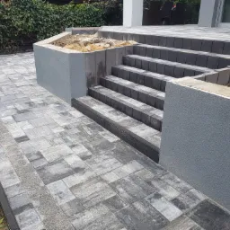 Ułożona kostka brukowa w odcieniach szarości tworząca taras i schody z podwyższonymi rabatami, widoczne niedokończone prace ziemne w obrębie rabat.