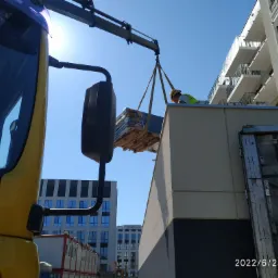 Żółta ciężarówka z dźwigiem podnosi paletę z materiałami budowlanymi na dach budynku, w tle nowoczesna architektura.