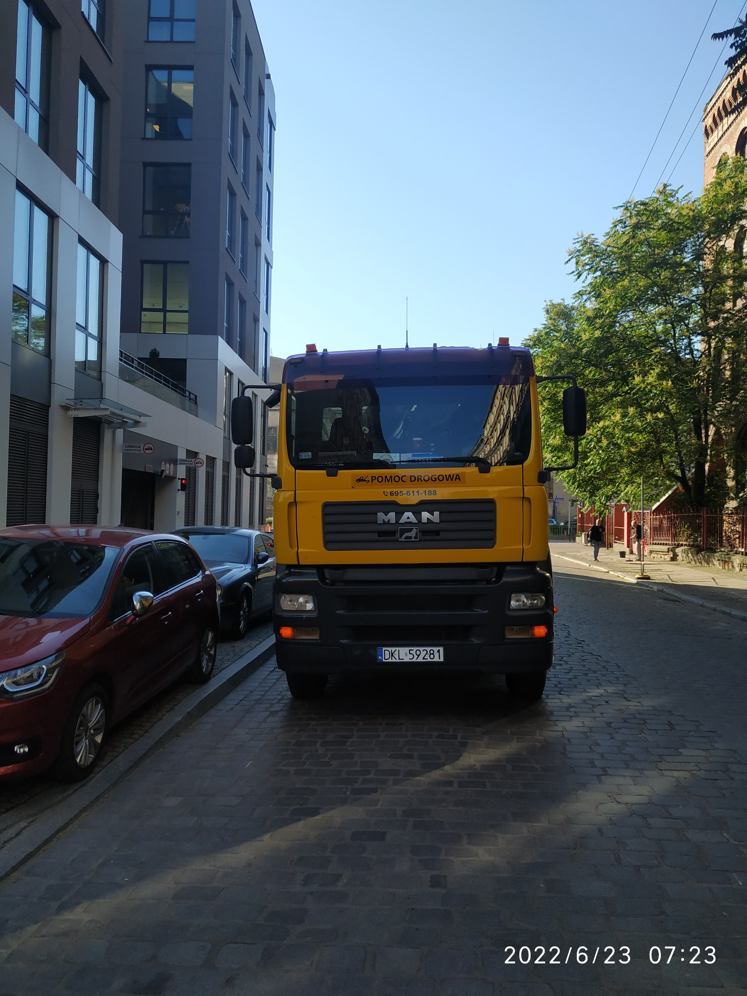 Żółty samochód ciężarowy MAN z napisem 'Pomoc Drogowa' zaparkowany na ulicy wyłożonej kostką brukową, w tle budynki biurowe i drzewa.