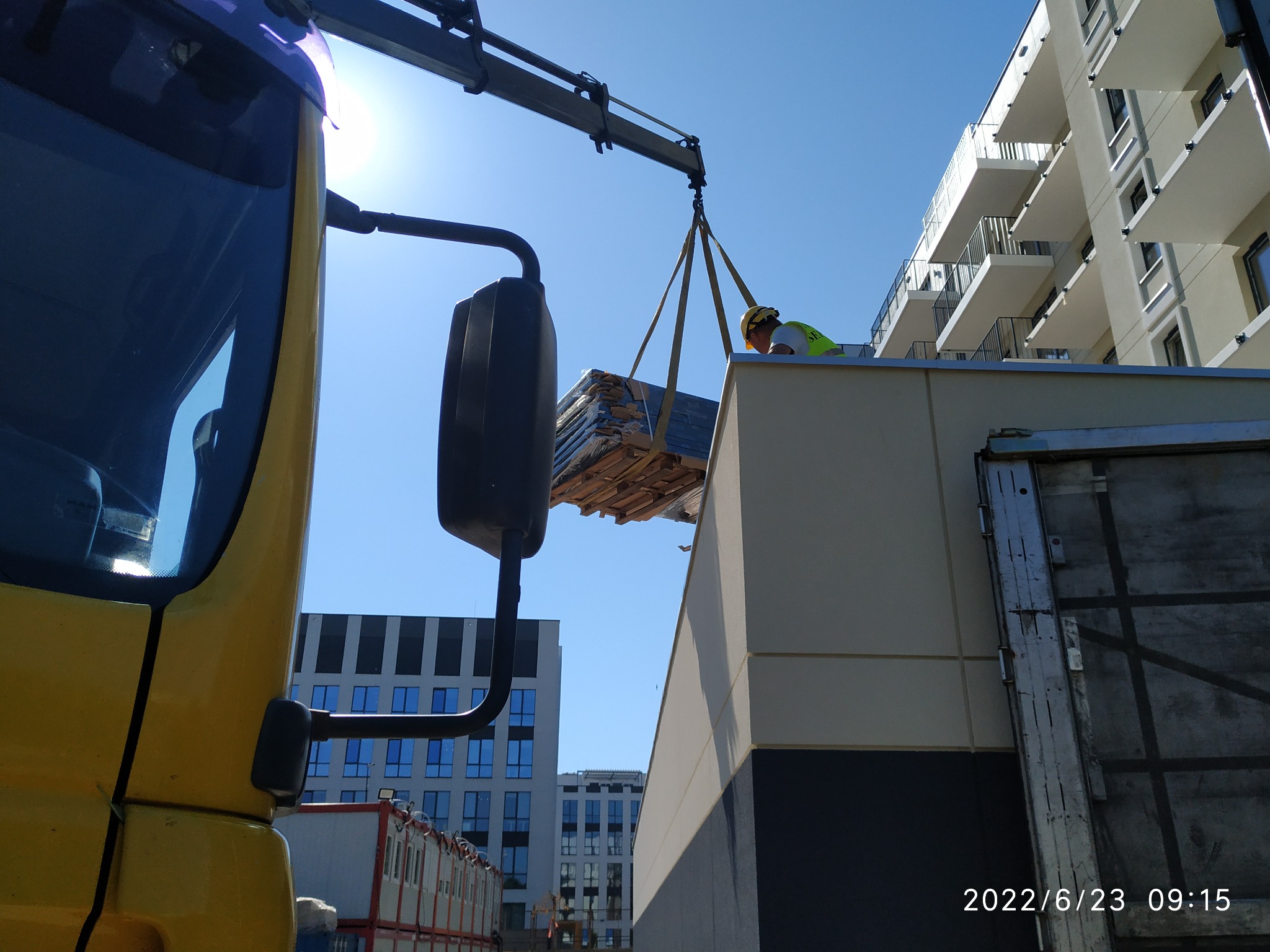 Żółta ciężarówka z dźwigiem podnosi paletę z materiałami budowlanymi na dach budynku, w tle nowoczesna architektura.