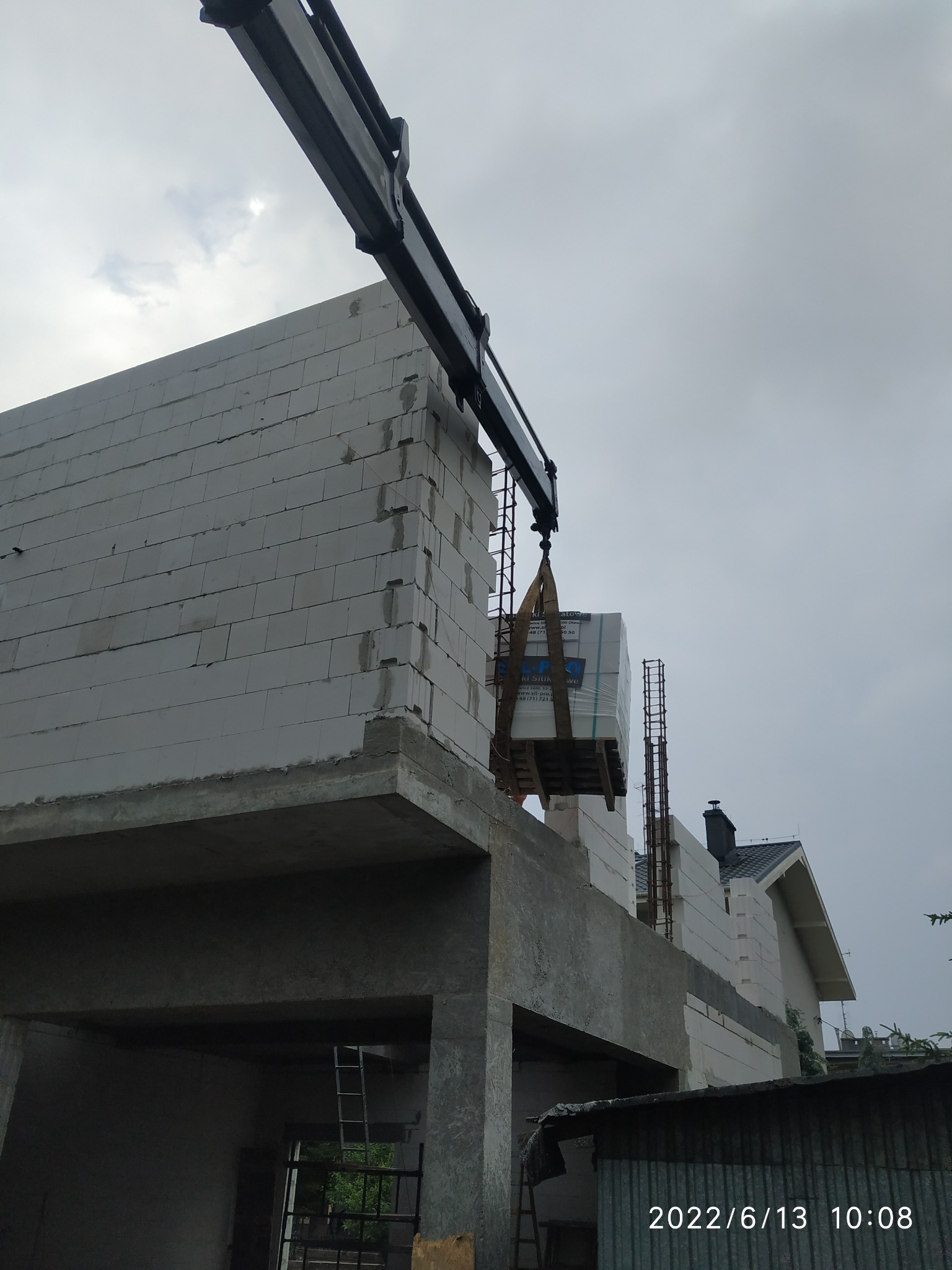 Żuraw budowlany podnosi paletę z bloczkami silikatowymi na budowę domu z białych pustaków, widoczne zbrojenia i data wykonania zdjęcia.