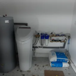 Szary bojler, biały zmiękczacz wody i instalacja wodna z filtrami, zaworami oraz worki z solą tabletkową w pomieszczeniu technicznym.