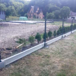 Panelowe ogrodzenie z podmurówką i słupkami, w tle domy i zieleń, widok z perspektywy trawnika.