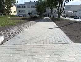 Szara, brukowana ścieżka prowadząca do budynku szpitala w słoneczny dzień. Widoczny trawnik i drzewa. Perspektywa z dołu, podkreślająca długość ścieżki.