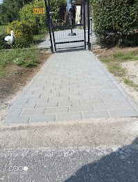 Szara kostka brukowa prowadząca do metalowej furtki, otoczona zielenią i krzewami. W tle widoczna biała dekoracja ogrodowa w kształcie ptaka.