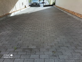 Szara kostka brukowa na podjeździe, widok z góry, z samochodem w tle za otwartą bramą. Minimalistyczny design, równa powierzchnia, delikatne chwasty w spoinach.