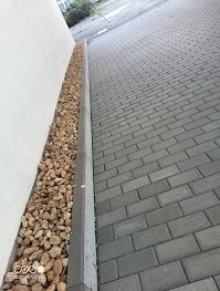 Szara kostka brukowa ułożona w jodełkę, oddzielona od białej ściany pasem jasnych kamieni ozdobnych. Widok z góry, lekko z ukosa.