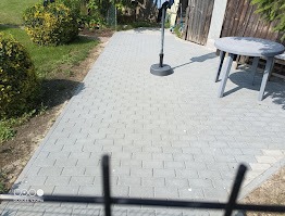 Szara kostka brukowa na podwórku, widok zza czarnego ogrodzenia. W tle trawnik, krzewy i stół z krzesłami. Słoneczny dzień.