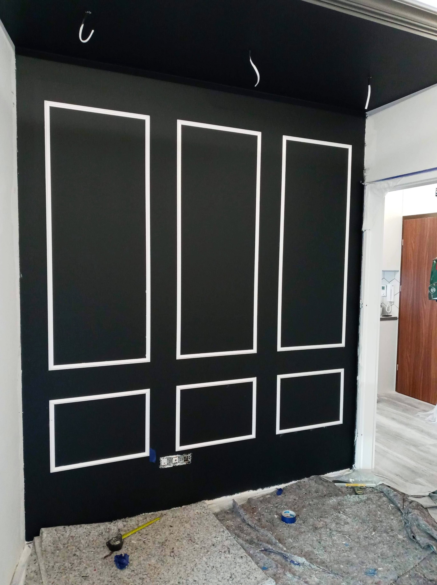 Czarna ściana z białymi, prostokątnymi listwami dekoracyjnymi w trakcie montażu, widoczne przygotowane podłoże, wystające przewody elektryczne i narzędzia pomiarowe.