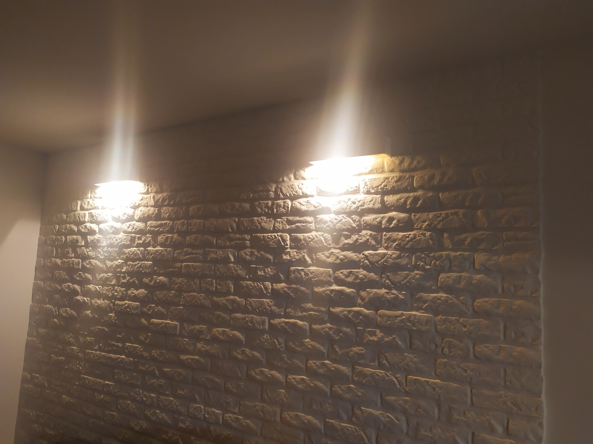 Ściana z dekoracyjnej cegły w Warszawie, oświetlona dwoma lampami dającymi ciepłe światło, tworzącymi grę cieni na fakturze cegieł.