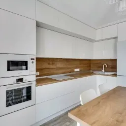 Jasna kuchnia z białymi, lakierowanymi frontami, drewnianym blatem i panelem ściennym, zabudowaną lodówką i piekarnikiem z mikrofalówką.