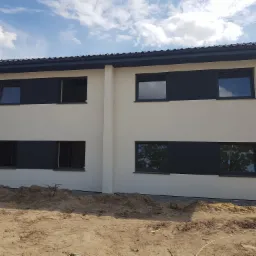 Biała elewacja dwukondygnacyjnego domu z czarnymi ramami okiennymi, widok na budynek z poziomu gruntu z widocznym piaskiem i elementami budowlanymi.