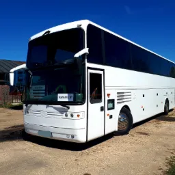 Biały, piętrowy autokar turystyczny zaparkowany na szutrowej nawierzchni przed budynkiem gospodarczym w słoneczny dzień.
