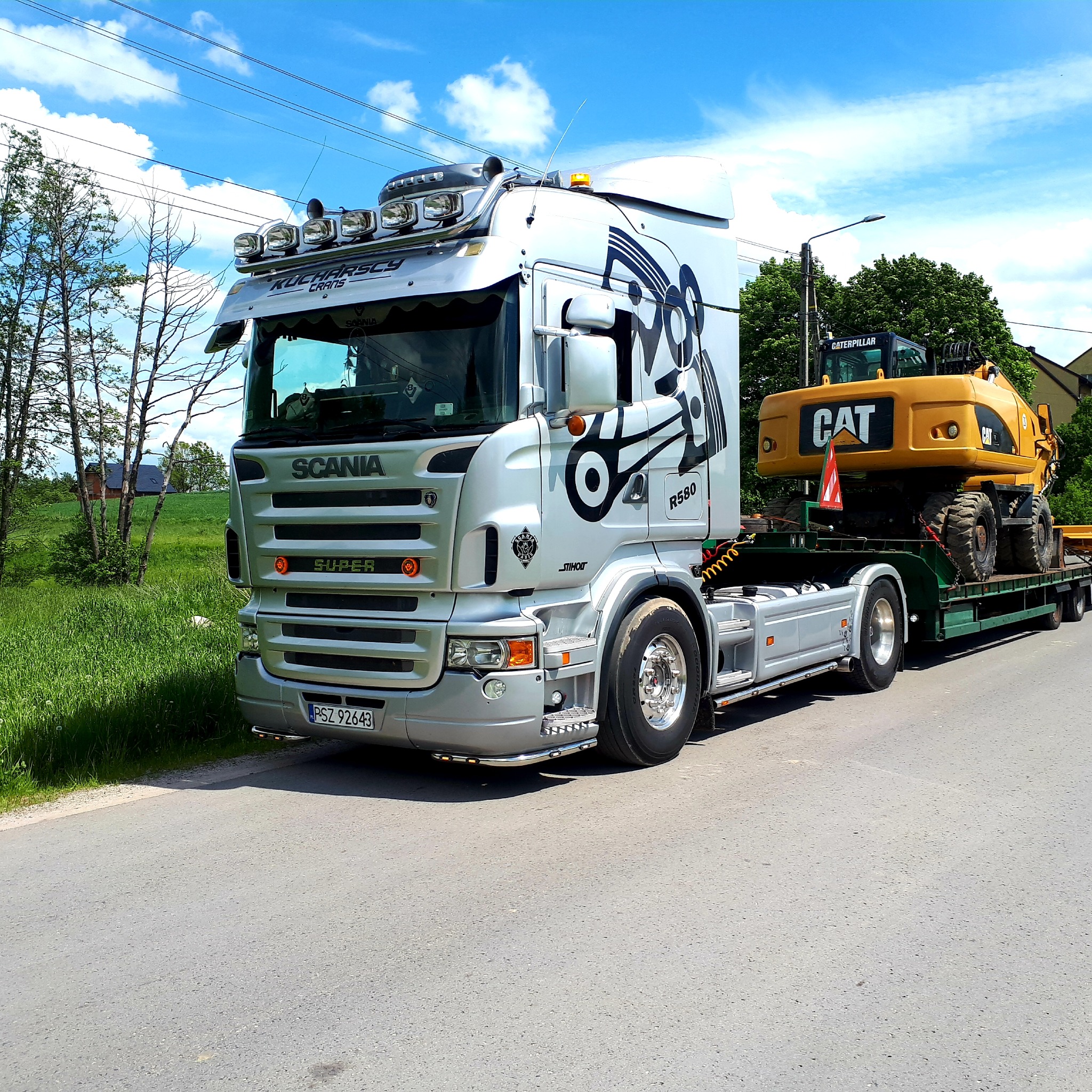 Srebrna ciężarówka Scania z czarnymi grafikami na kabinie przewozi żółtą koparkę Caterpillar na niskopodwoziowej platformie, widok z boku na tle zieleni i błękitnego nieba.