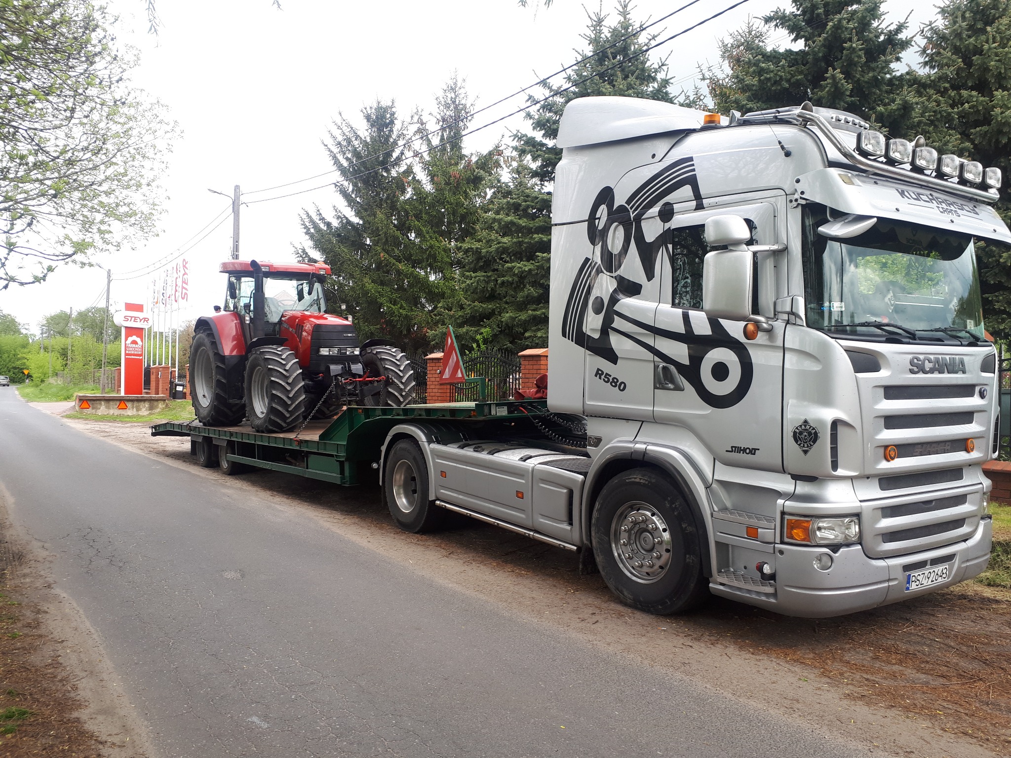 Laweta Scania przewozi czerwony traktor Steyr na zielonej platformie, widoczny znak firmowy Steyr w tle.
