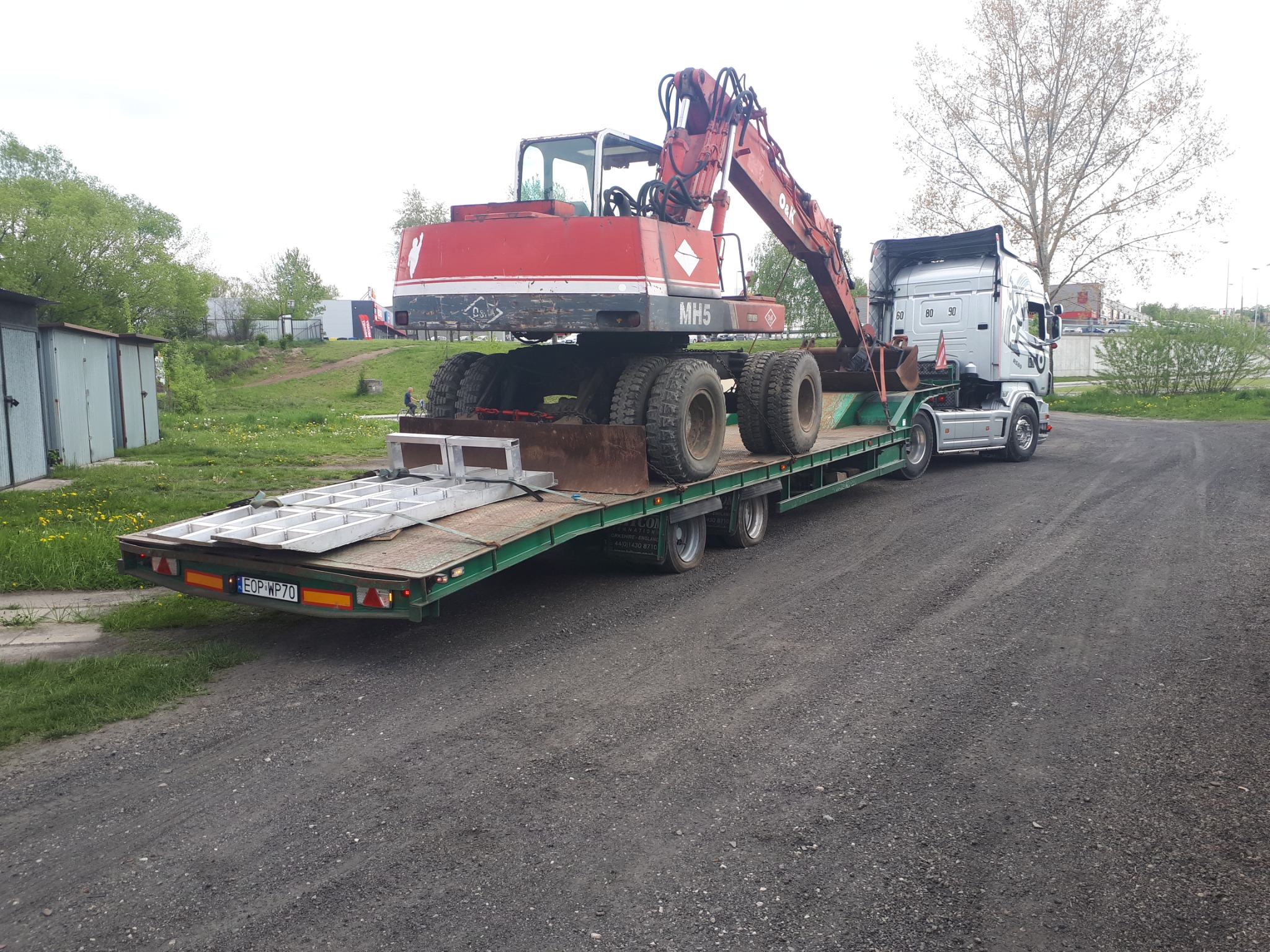 Ciężarówka z naczepą niskopodwoziową przewożąca czerwoną koparkę kołową MH5 na żwirowej drodze, widoczne garaże w tle.