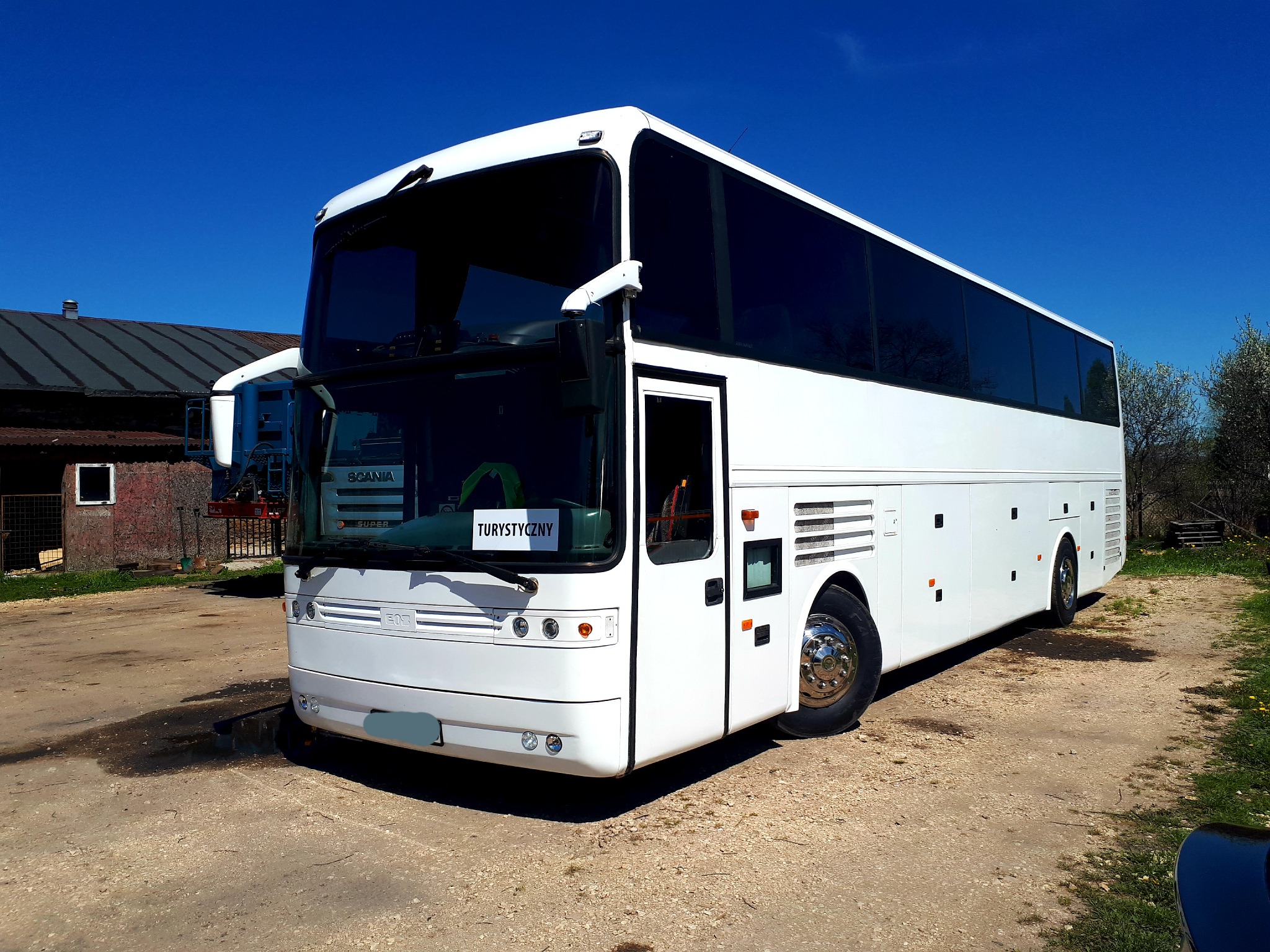 Biały, piętrowy autokar turystyczny zaparkowany na szutrowej nawierzchni przed budynkiem gospodarczym w słoneczny dzień.