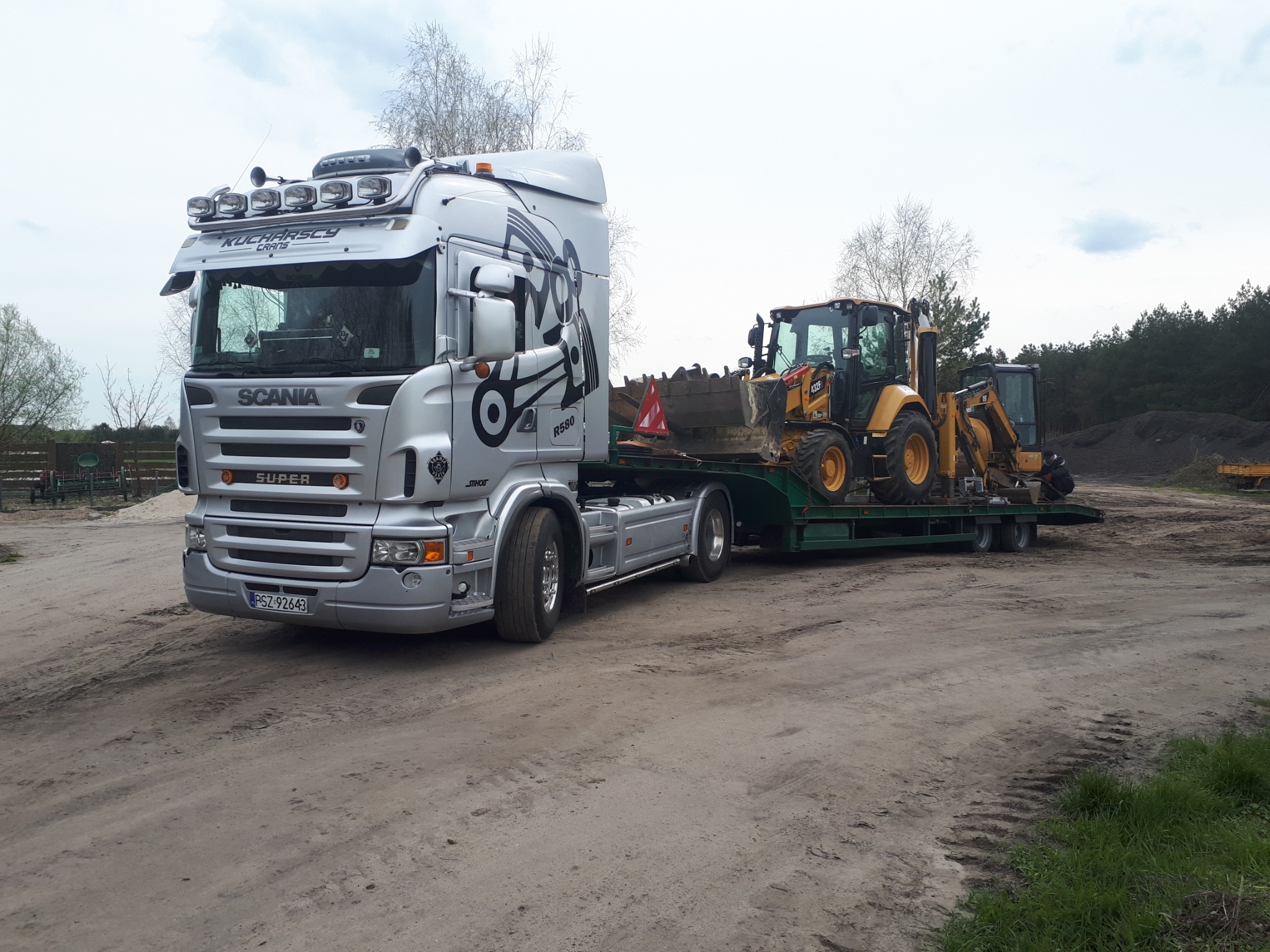 Srebrna ciężarówka SCANIA z naczepą niskopodwoziową przewożąca dwie żółte koparko-ładowarki na szutrowej drodze.