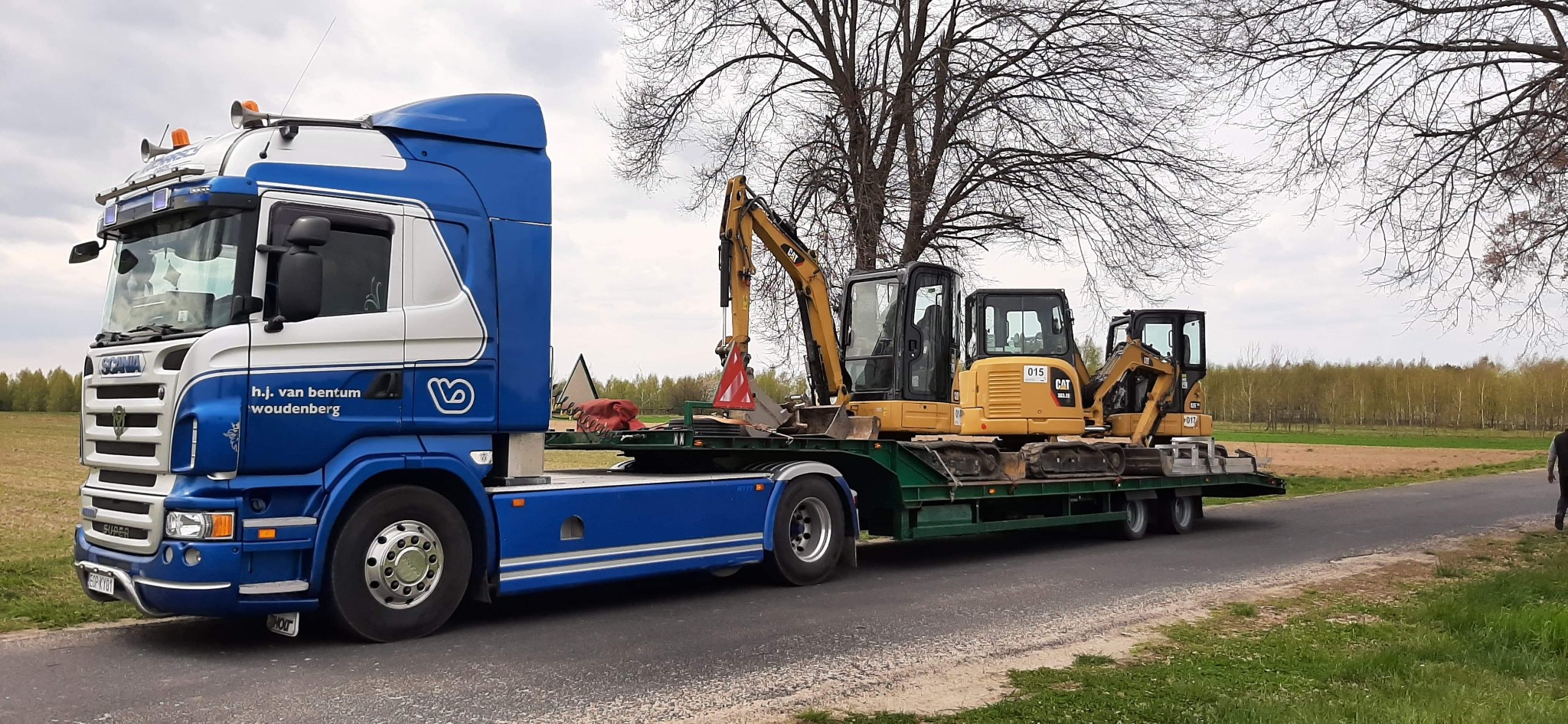 Niebiesko-biała ciężarówka Scania firmy h.j. van bentum woudenberg przewozi na niskopodwoziowej naczepie trzy żółte minikoparki CAT, na tle pola i drzew.