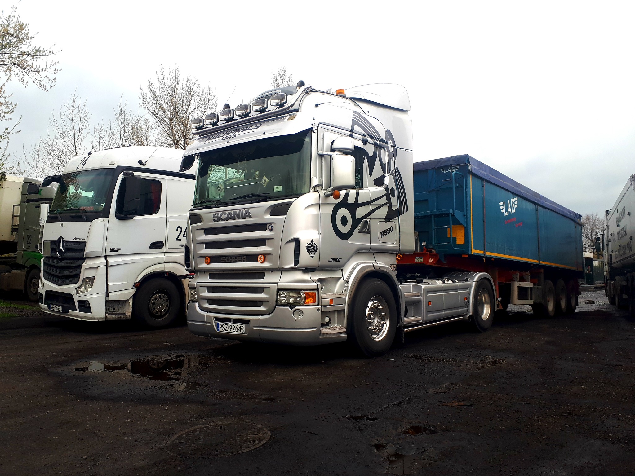 Srebrny ciągnik siodłowy Scania R580 z niebieską naczepą LAG na błotnistym placu, obok biały Mercedes Actros z numerem 24, widoczne drzewa bez liści w tle.