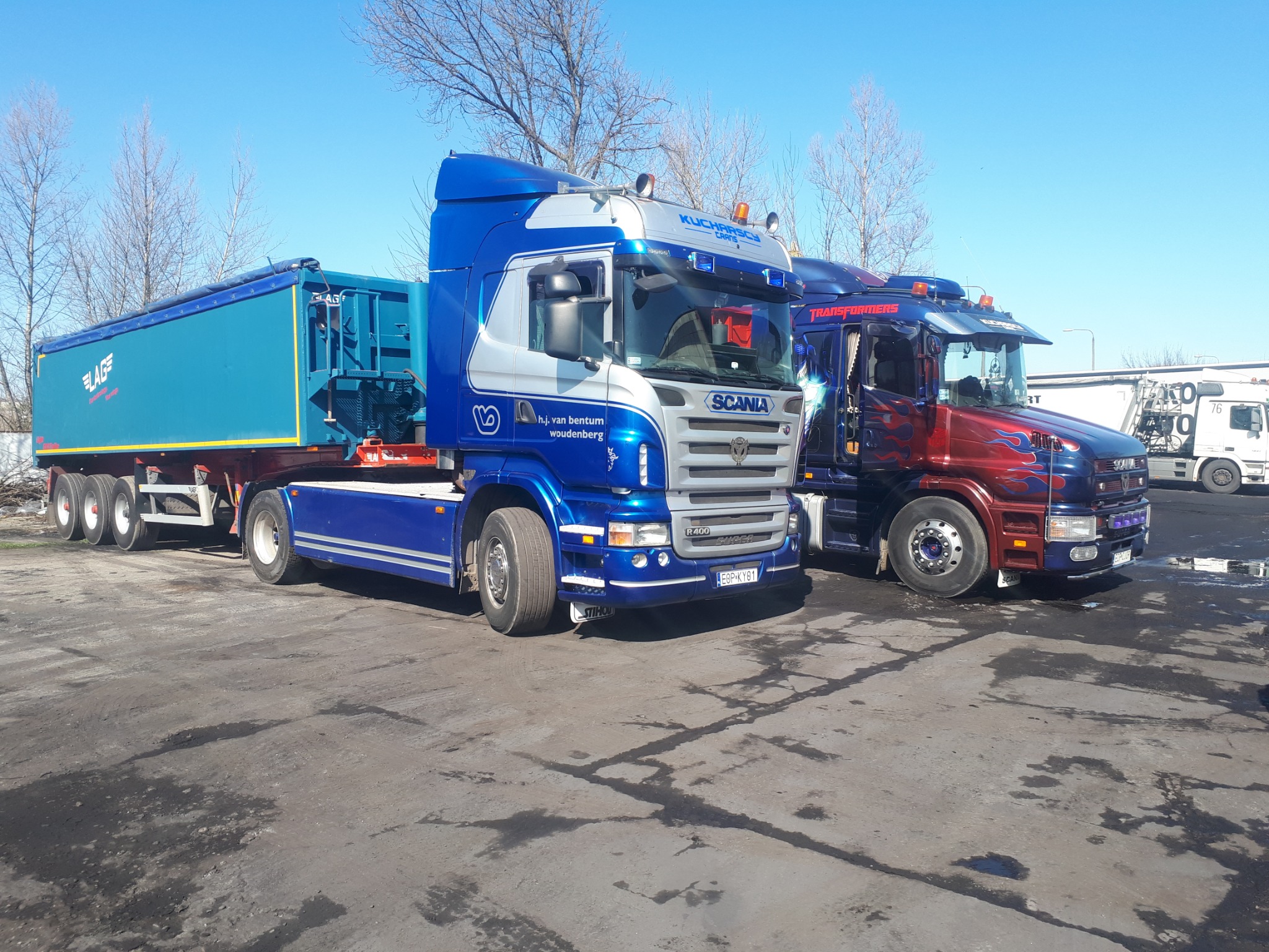 Dwa samochody ciężarowe Scania zaparkowane obok siebie na placu. Jeden z nich to niebiesko-srebrny ciągnik z naczepą LAG, drugi ma motyw Transformers z płomieniami. Widoczne logo firm. Słoneczny...