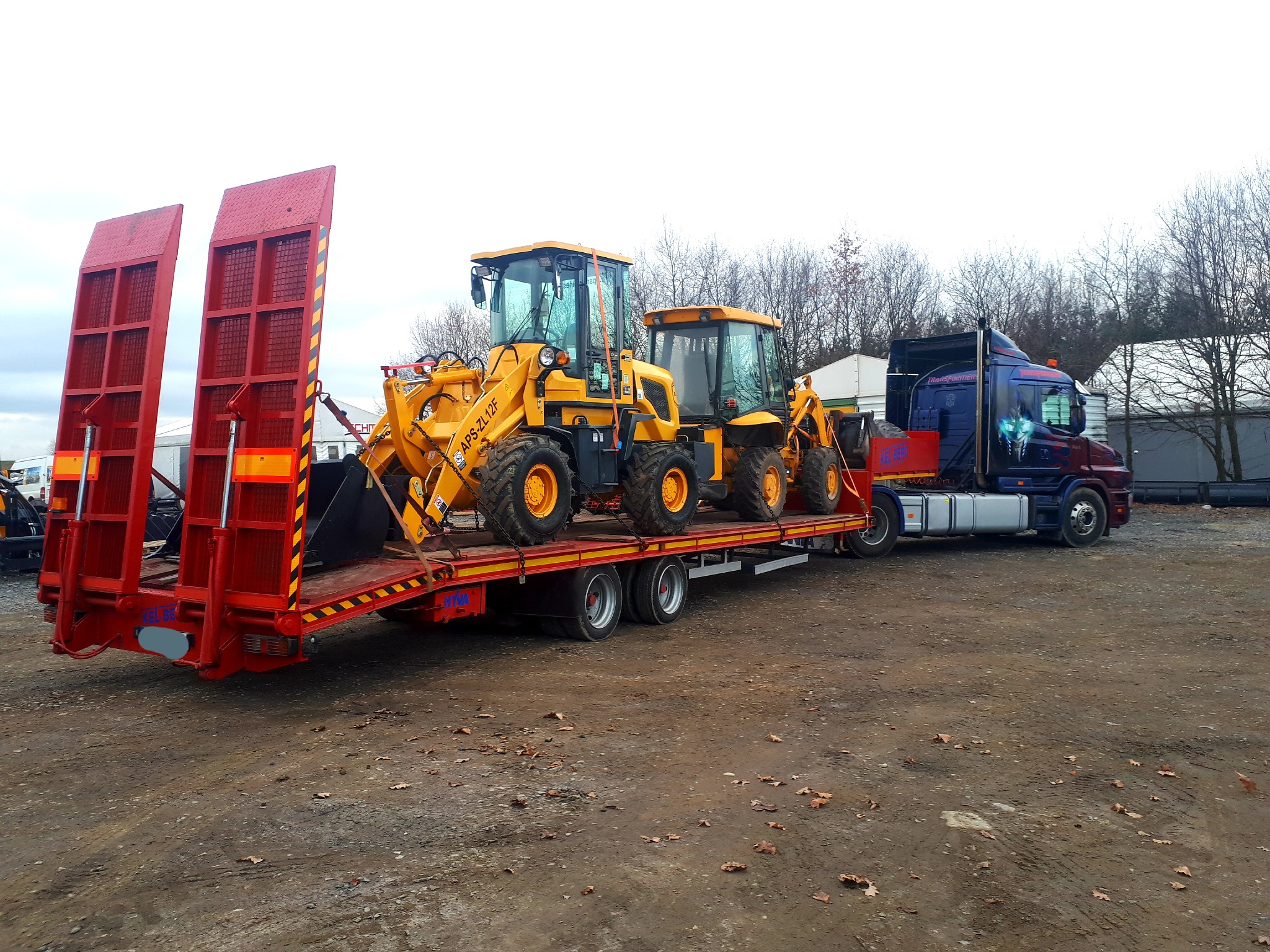 Ciężarówka z naczepą typu laweta przewożąca dwie żółte ładowarki kołowe z oznaczeniem APS-ZL12F na tle drzew i budynków.