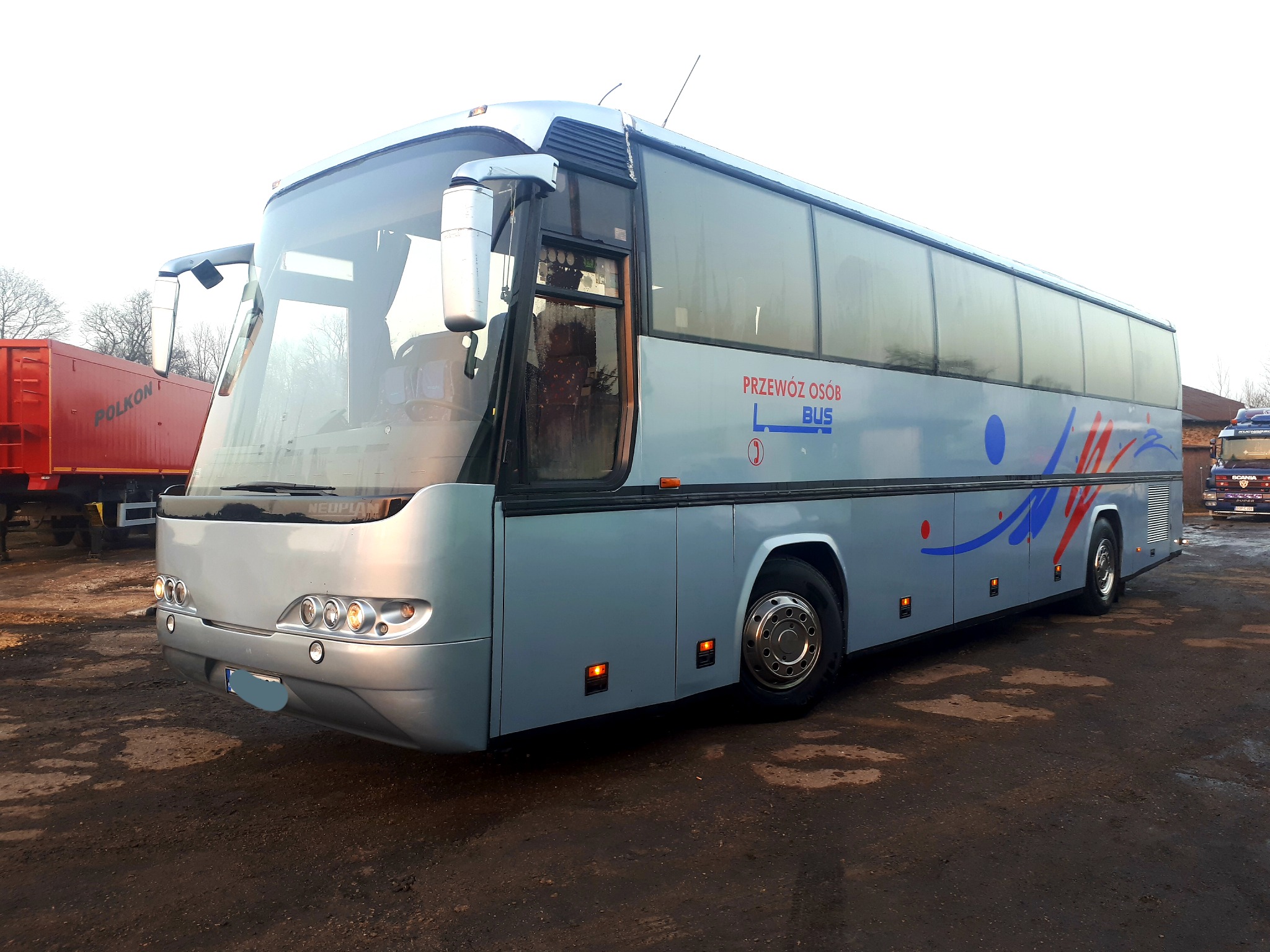 Srebrny autobus Neoplan z napisem 'Przewóz Osób BUS' na boku, stojący na błotnistym placu, z czerwonym kontenerem i innymi pojazdami w tle.