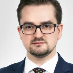 Portret mężczyzny w okularach, garniturze i wzorzystym krawacie na jasnym tle.