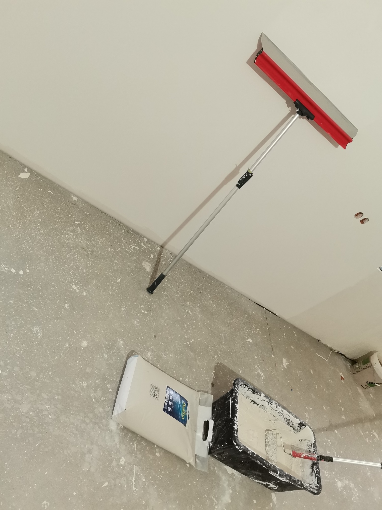 Aplikator z długim uchwytem rozprowadza gładź na suficie; obok stoi wiadro z gładzią i wałek; na podłodze leży worek z zaprawą.