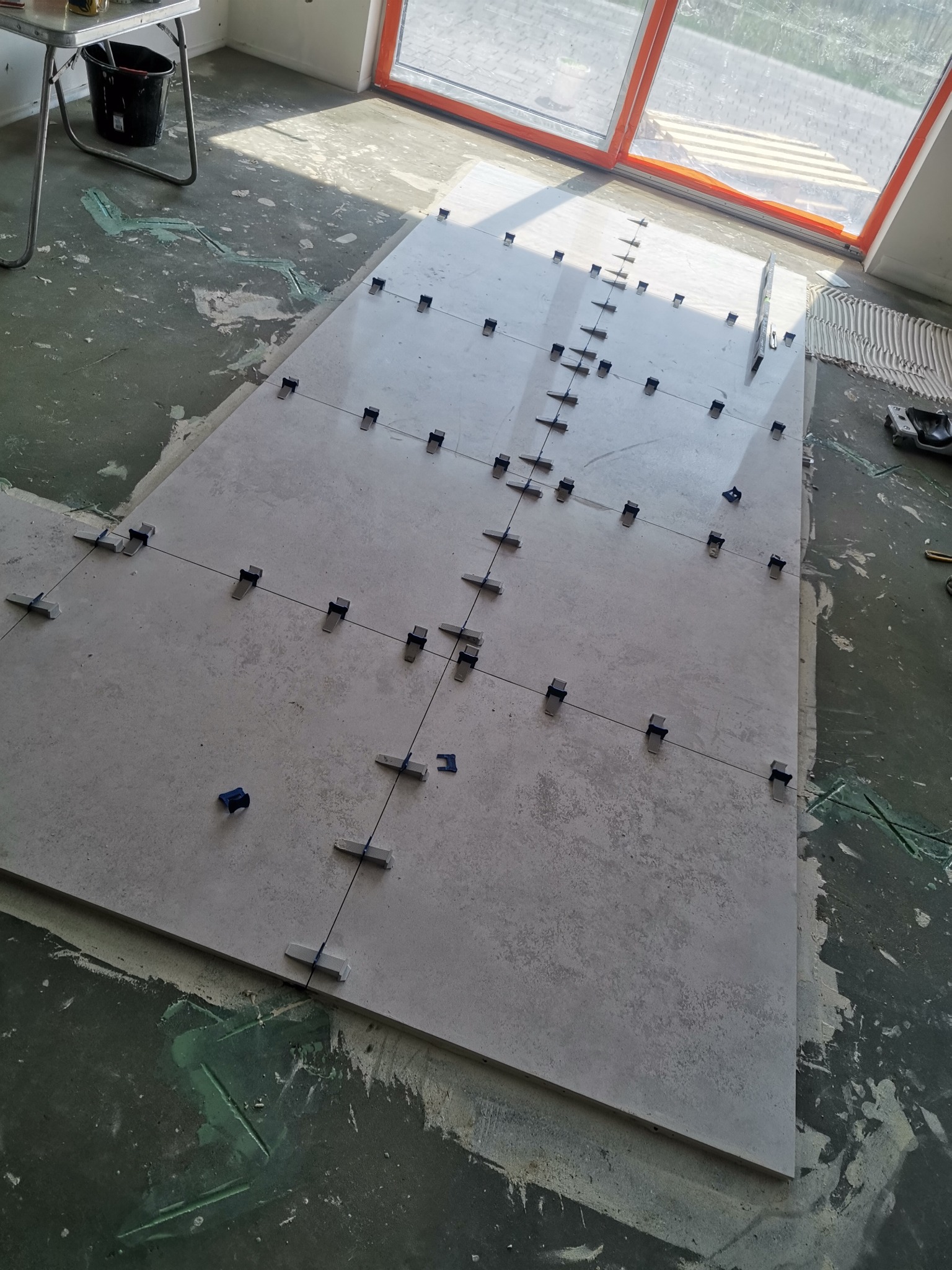 Układanie jasnych, kwadratowych płytek podłogowych z użyciem klipsów poziomujących i sznurka krzyżowego w trakcie remontu, widoczne ślady kleju na podłożu.