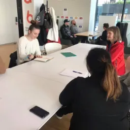 Grupa osób w jasnym biurze, siedząca przy dużym białym stole podczas spotkania, notująca w notatnikach, z telefonem leżącym na stole, w tle widać osoby siedzące przy innym stole i dużą szybę...