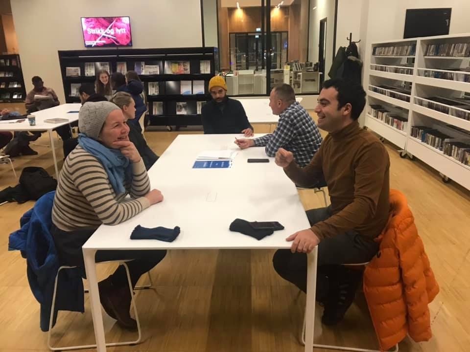 Grupa osób siedzi przy białych stołach w jasnym pomieszczeniu, prawdopodobnie bibliotece lub sali konferencyjnej, dyskutując i patrząc na dokumenty leżące na stole. Jeden mężczyzna w brązowym...