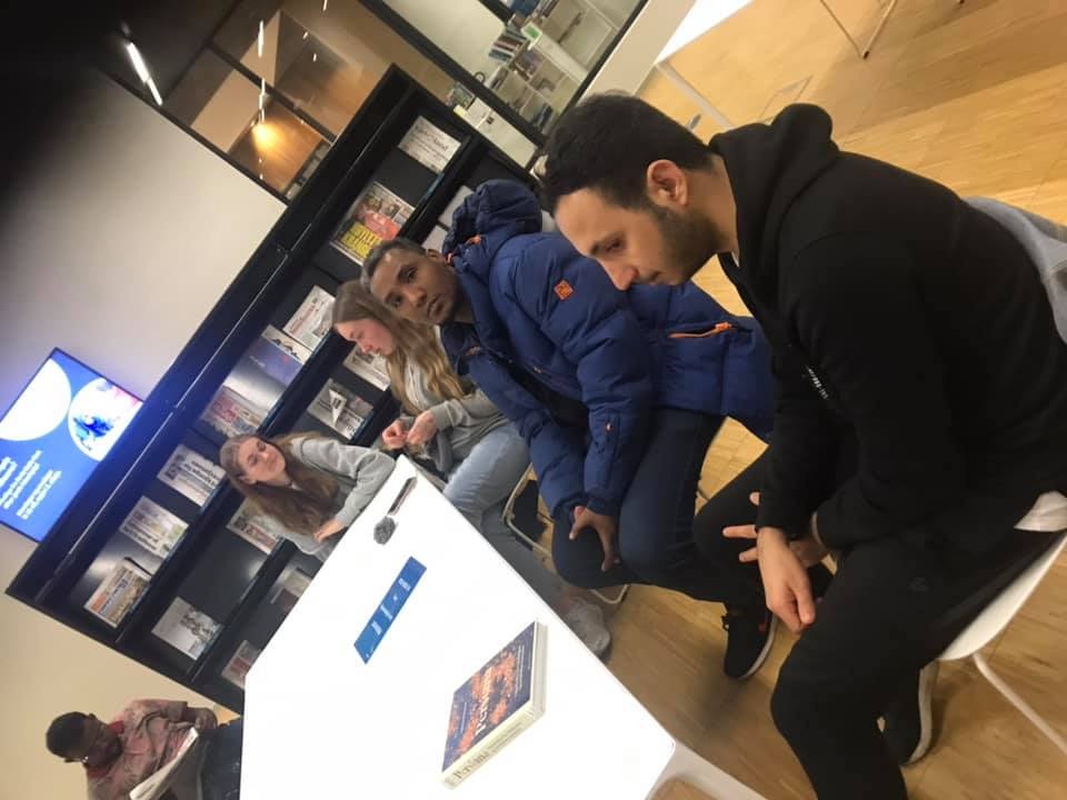 Grupa młodych osób siedzi przy białym stole w pomieszczeniu z regałem pełnym gazet i ulotek w tle, możliwe, że omawiają warunki umowy lub oferty finansowej.