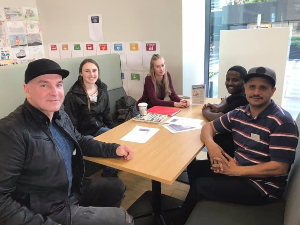 Grupa pięciu osób siedzi przy drewnianym stole w jasnym pomieszczeniu, otoczona dokumentami i kubkiem, sugerując spotkanie lub konsultację finansową.