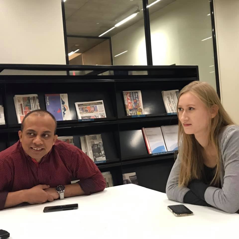 Dwie osoby siedzą przy białym stole w nowoczesnym biurze z regałem pełnym gazet i książek w tle, prawdopodobnie podczas rozmowy lub spotkania.