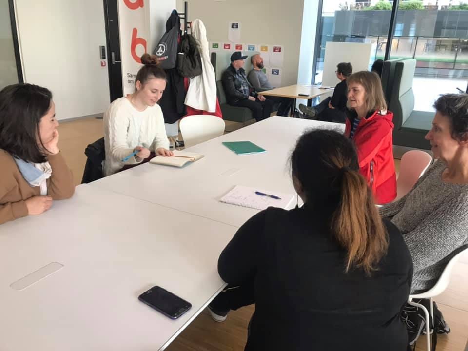 Grupa osób w jasnym biurze, siedząca przy dużym białym stole podczas spotkania, notująca w notatnikach, z telefonem leżącym na stole, w tle widać osoby siedzące przy innym stole i dużą szybę...