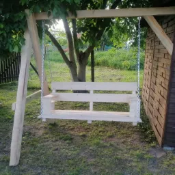 Surowa, drewniana huśtawka ogrodowa zawieszona na łańcuchach, w tle zielony ogród i drewniana altana, widoczne niedoskonałości obróbki drewna.